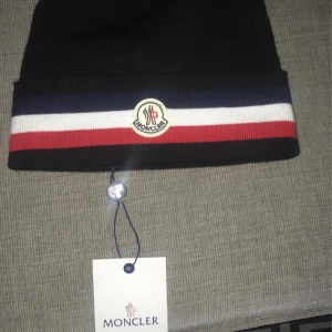 Moncler Tricolor beanie - Snygg Moncler mössa i mörkblått med breda ränder i vitt och rött. Klassisk Moncler-logga framtill. Mössan är stickad och har en clean, sportig look som passar perfekt till streetwear. Kommer med originalförpackning och taggar. Helt oanvänd kommer med box, kvitto, taggar och moncler påse