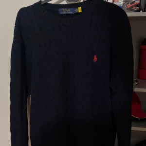 Mörkblå stickad tröja Polo Ralph Lauren - Kabelstickad mörkblå tröja från Polo Ralph Lauren med klassisk rund halsringning och röd broderad logga på bröstet. Långärmad och i mjuk bomull, perfekt för lager-på-lager under kyliga dagar.