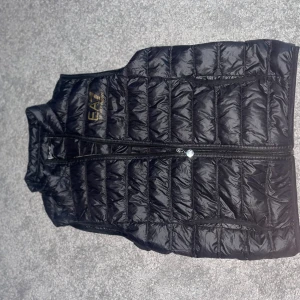 EA7 Emporio Armani väst - Hej säljer nu min Emporio Armani väst då den är för liten, de finns inga skador på västen.  Storlek S har jag för mig. De finns två ytter fickor och två inne fickor. Den är svart med silver dragkedja och guld detalj som huvud märke, de är bara skrivs på om frågor 😊