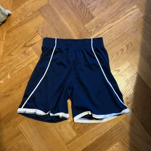 Blå sportshorts med vita detaljer - Säljer ett par marinblå sportshorts med vita ränder längs sidorna och vit kant nertill. Shortsen har elastisk midja och är tillverkade i ett lätt och svalt syntetmaterial, perfekta för träning eller sportiga aktiviteter.