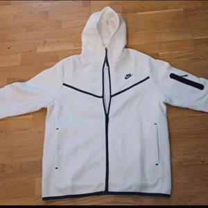 Vit Nike vindjacka med huva - Snygg vit tracksuit från Nike med svart dragkedja och detaljer. Jackan har huva, lång ärm och två fickor framtill. Svart Nike-logga på bröstet och cool svart detalj på ena ärmen. Perfekt för dig som gillar sportig och clean stil.