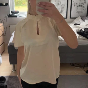Champagnefärgad blus  - Säljer en elegant champagnefärgad blus från b.young i storlek 34. Blusen har korta ärmar, hög krage och en snygg v-ringad öppning framtill med liten knappdetalj. Materialet är glansigt och känns lyxigt mot huden. Perfekt för dig som gillar stilrena och trendiga plagg.