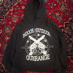 Vicinity Hoodie - Svart hoodie från Vicinity med stor vit print framtill med texten 'INDOOR/OUTDOOR GUNRANGE' och motiv av två korsade pistoler. Klassisk huva och känguruficka. Perfekt för dig som gillar streetwear och statement-plagg.