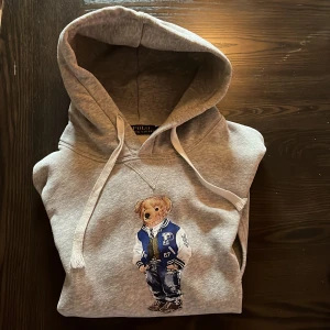 Grå Polo Bear hoodie från Ralph Lauren - Säljer en grå hoodie från Ralph Lauren med ikoniska Polo Bear-trycket på bröstet. Tröjan har huva med snörning,. Tillverkad i mjuk bomullsmix, perfekt för chill dagar. Klassisk och stilren hoodie. Skriv gärna om ni undrar något 