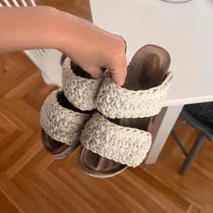 Krämvita sandaler med flätade band - Säljer ett par snygga sandaler med två breda, flätade band i beige textil. Sandalerna har öppen tå och platt sula med en naturlig träfärgad innersula. Perfekta för sommarens alla dagar och ger en chill boho-vibe till din outfit.