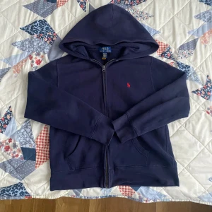 Mörkblå hoodie från Polo Ralph Lauren - Mörkblå hoodie från Polo Ralph Lauren med dragkedja framtill och klassisk röd broderad logga på bröstet. Tröjan har huva, kängurufickor och är tillverkad i mjuk bomull. Perfekt för en chill och stilren look.