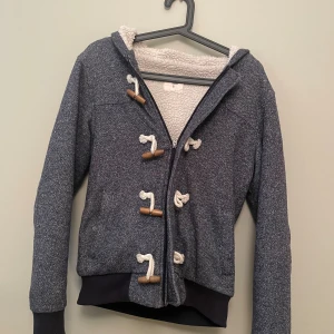 Mörkgrå cardigan -  mörkgrå cardigan med vit fluffig insida och huva. Jackan har snygga träknappar med grova snören och dragkedja. Muddar vid ärmslut och nederkant ger en skön passform. Perfekt för kyliga dagar och en chill streetstyle.