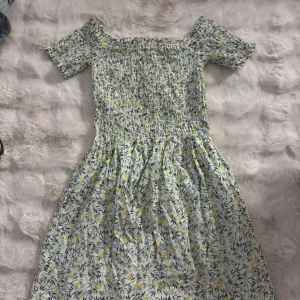 Blommig offshoulder sommarklänning - Söt blommig klänning med offshoulder och korta ärmar. Mönstret har små gula och gröna blommor på vit botten. Smockad överdel och luftig kjol ger en skön passform. Perfekt för varma dagar och har en fräsch, ungdomlig vibe.