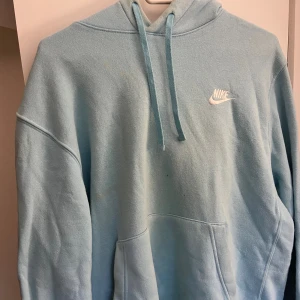 Ljusblå hoodie från Nike - Säljer en ljusblå hoodie från Nike med klassisk logga broderad på bröstet. Tröjan har huva med snörning och en stor magficka. Perfekt för dig som gillar en avslappnad och sportig stil.