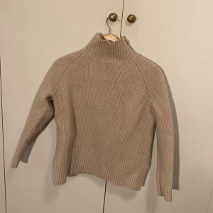 Beige ribbad polotröja  - Säljer en beige ribbstickad polotröja med hög krage. Tröjan är i mjuk ull och har lång ärm och klassisk passform. Perfekt för lager på lager under kyliga dagar.