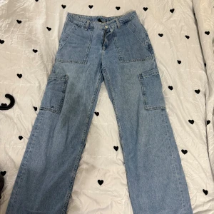 Blå cargopants i jeansmaterial - Säljer ett par blå cargopants i jeansmaterial med breda ben och stora fickor på sidorna. Byxorna har hög midja och klassisk knappstängning framtill. Perfekt för dig som gillar streetstyle och vill ha en cool look.