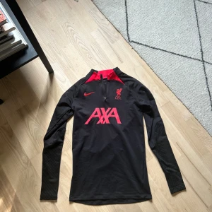 [SÅLD] Liverpool långärmad Nike Dri-Fit XS - Liverpool FC tränings-tröja från Nike i slim fit-modell, tillverkad i Dri-Fit material som andas. Svart bas med röda detaljer vid kragen och tryck på bröstet, inklusive AXA-logga och klubbmärke. Lång dragkedja framtill och diskret mönster på ärmarna.