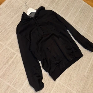 Svart Hugo Boss hoodie med snörning, storlek M - Svart Hugo boss hoodie i storlek M med snörning vid huvan och en stor magficka framtill. Tröjan har en enkel och clean look med ton-i-ton broderad text på bröstet. Perfekt för dig som gillar snygga enkla hoodies med stil och vill ha en bekväm och stilren tröja. Passar M-S