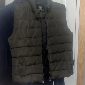 Mörkgrön dunväst från Stone Island - Snygg mörkgrön dunväst från Stone Island med quiltad design och dragkedja framtill. Västen har två praktiska fickor på sidorna och en hög krage som ger extra stilpoäng. Perfekt att slänga över en hoodie för en cool look.