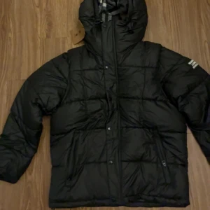 Svart Burberry pufferjacka med huva, avtagbara ärmar - Svart pufferjacka från Burberry med avtagbara ärmar, vilket gör att den kan användas som väst. Jackan har huva med klassiskt rutigt Burberry-foder, dragkedja och knappar framtill samt fickor med dragkedja. Märkeslogga 'Burberry London England' på ärmen.