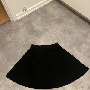 Svart kortkjol med A-linjeform - Säljer en svart kortkjol med snygg A-linjeformad siluett. Kjolen har en enkel och stilren design utan synliga detaljer eller mönster, vilket gör den lätt att matcha med olika toppar. Materialet känns mjukt och följsamt mot kroppen. Perfekt för dig som gillar minimalistisk stil. En aning nopprig.