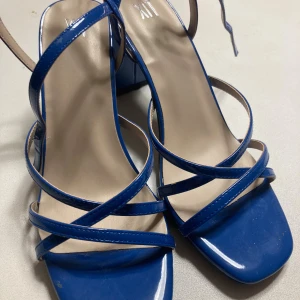 Blå sandaler med klack från XIT - NYA - Snygga blå sandaler från XIT med flera smala remmar och fyrkantig tå. Klackarna är stabila och ger extra höjd. Remmarna har en glansig finish och spänne vid ankeln. Perfekt för dig som vill sticka ut med färg och stil.
