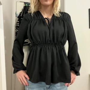 Svart blus med knyt och v-ringning - Säljer en svart blus med v-ringning och knyt framtill. Blusen har långärmade, lätt puffiga ärmar och markerad midja med resår som ger en snygg siluett. Materialet är mjukt och har ett lätt glansigt utseende. Perfekt till jeans eller kjol för en cool look. Märker är Wera och är i storlek 36. 