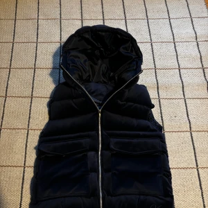 Svart dunväst med huva och fickor - Svart dunväst från vendier med stor huva och två rejäla fickor framtill. Västen har en hel dragkedja och quiltad design som ger en cool look. Insidan är fodrad med blankt syntetmaterial. Perfekt för lager-på-lager under höst och vinter. Pris kan diskuteras tar även byten hör av dig vid minsta fundering