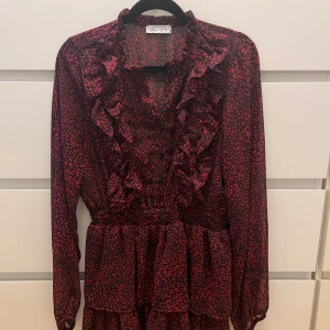 Röd leopardmönstrad klänning DeFacto - Snygg kort klänning från DeFacto med rött leopardmönster på svart botten. Klänningen har volangdetaljer längs halsringningen och framtill, samt långärmade ärmar och markerad midja. Klännningen är storlek 40 men passar som en xs skulle jag säga.