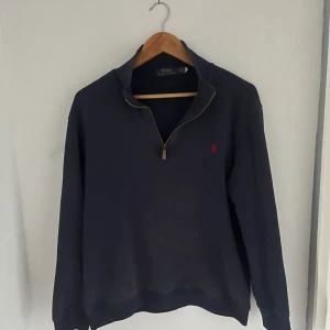 Mörkblå half zip tröja Polo Ralph Lauren - Mörkblå långärmad tröja från Polo Ralph Lauren med half zip och klassisk röd logobrodyr på bröstet. Tröjan har en ståkrage och är tillverkad i mjuk bomull, perfekt för chill dagar. Snygg och enkel att matcha med jeans eller sweatpants.