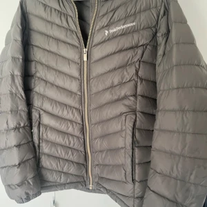 Grå dunjacka Peak Performance M - Snygg grå dunjacka från Peak Performance med huva och guldfärgad dragkedja. Jackan har ett quiltat mönster och är tillverkad i lätt polyester med dunfyllning. Perfekt för kalla dagar och har en stilren look med logga på bröstet.