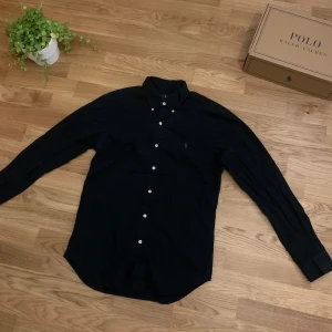 Svart slim fit skjorta från Ralph Lauren - Svart skjorta från Ralph Lauren i slim fit-modell, klassisk button-down krage och vita knappar framtill. Skjortan har lång ärm och är tillverkad i mjuk bomull. Diskret lila Polo-logga broderad på bröstet för en clean look.