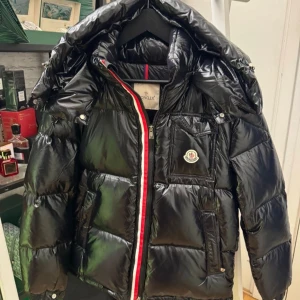 Svart pufferjacka från Moncler - Säljer en svart pufferjacka från Moncler med glansig finish och huva. Jackan har en rödvit detalj vid dragkedjan, flera fickor och Moncler-logga på bröstet. Perfekt för kalla dagar och har en riktigt snygg streetstil. En riktigt snygg jacka som är perfekt för en kall vinterdag. Hör av er i dm vid minsta lilla. 2799 är priset men de är ej hugget i sten. Tack i förväg. Har alla storlekar…