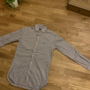 Rutig skjorta från Polo Ralph Lauren - Säljer en klassisk rutig skjorta från Polo Ralph Lauren i bomull. Skjortan har en snygg mix av rosa, grön och blå toner, broderad logga på bröstet och knappar framtill. Perfekt för dig som gillar preppy stil och vill ha något som sticker ut lite extra.