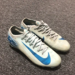 Nike fotbollsskor i vitt och blått - Säljer ett par Nike fotbollsskor i vit och blå färg med snörning och rund tå. Skorna har en syntetisk ovandel med blå Nike-logga och mönstrade detaljer på hälen. Sulan har dobbar för bra grepp på planen. Perfekt för dig som vill ha stil på fotbollsplanen.