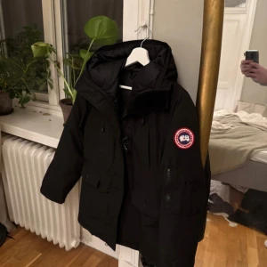 Svart dunjacka från Canada Goose - OBS- hål på ena fickan, går att sy” Svart dunjacka från Canada Goose med huva och klassisk logga på ärmen. Jackan har flera fickor framtill, dragkedja och knäppning. Perfekt för kalla vinterdagar och har en stilren look som funkar till det mesta.