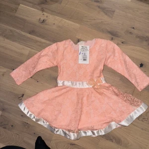 Rosa klänning, storlek 1-2 år Rosa spetsklänning med rosett - Söt rosa klänning med lång ärm och vit banddetalj. Perfekt för festliga tillfällen. Supersöt rosa kortklänning från S&V Vitex med långärmade spetsdetaljer och en vit satinband i midjan. Klänningen har en dekorativ spetsrosett på sidan och ett glansigt band längs nederkanten. Perfekt för dig som gillar romantisk stil. 🎁 🎄 🎉 