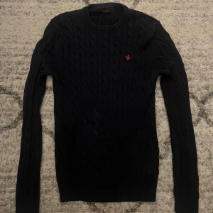 Marinblå kabelstickad tröja Morris M - Marinblå  kavelstickad tröja från Morris i storlek M. Tröjan har rund hals, lång ärm och ett rött broderat emblem på bröstet. Mjuk och skön kvalitet med klassisk ribbad kant vid hals, ärmslut och nederkant. Perfekt för dig som gillar stilrena och tidlösa plagg.