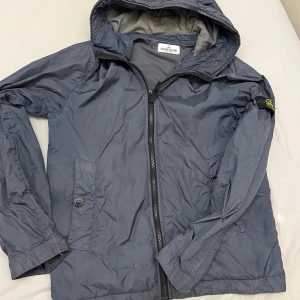 Blå vindjacka från Stone Island Junior - Snygg blå vindjacka från Stone Island Junior med huva och klassisk logga på ärmen. Jackan har dragkedja framtill, två fickor med knapp och är tillverkad i lätt polyester. Perfekt för dig som gillar streetwear och vill ha något stilrent.