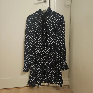 Prickig kortklänning från Bik Bok - Supersöt svart kortklänning från Bik Bok med vita prickar och volang i nederkant. Klänningen har långärmade puffärmar, hög hals med knytband och en flowy passform. Perfekt för dig som gillar retrovibbar och lekfull stil.