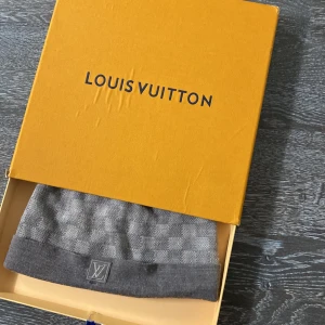 Grå Louis Vuitton mössa i ull - Snygg grå mössa från Louis Vuitton med klassiskt rutigt mönster och LV-logga framtill. Tillverkad i mjuk ullblandning som håller dig varm och stilren. Perfekt accessoar för att lyfta din vinterlook.