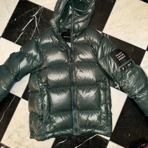 Mörkgrön Peak Performance pufferjacka - Snygg mörkgrön pufferjacka från Peak Performance med huva och stor patch på ärmen. Jackan har en glansig finish, dragkedja framtill och två stora fickor. Perfekt för kalla dagar och har en modern, boxig passform. Mycket bra skick!