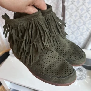 Olivgröna boots med fransar, stl 38 - Olivgröna boots i mocka med perforerat mönster framtill och fransar runt skaftet. Skorna har dragkedja baktill för enkel på- och avtagning samt platt sula. Perfekt för dig som gillar boho-stil och vill ha något unikt.