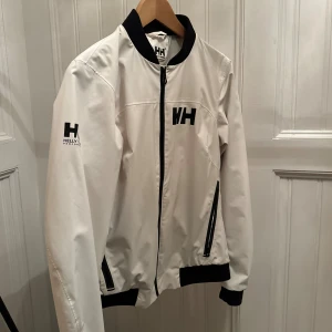 Vit vindjacka från Helly Hansen L - Snygg vit vindjacka från Helly Hansen med svarta detaljer och logga på bröstet och ärmen. Jackan har dragkedja framtill, svarta muddar och krage, samt två fickor med svarta dragkedjor. Perfekt för dig som gillar stilren och sportig look.