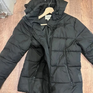 Svart pufferjacka från KappAhl - Svart pufferjacka från KappAhl med huva och två dragkedjeförsedda fickor. Jackan har quiltad design och Urban District-märke på ärmen. Perfekt för kalla dagar och har en snygg, enkel look som funkar till det mesta.