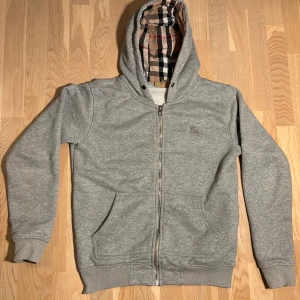 Grå hoodie från Burberry med dragkedja - Grå hoodie från Burberry med klassiskt rutigt foder i huvan. Tröjan har dragkedja framtill, två fickor och ribbade muddar. Tillverkad i mjuk bomullsmix och fodrad med mysig fleece på insidan. Perfekt för kyliga dagar.