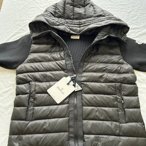 Svart Moncler cardigan  - Svart Moncler cardigan med quiltad framsida, stickade svarta  och ärmar, samt en stor huva. Västen har två fickor med tryckknappar och dragkedja framtill. Moncler-logga på vänster ärm och snygga detaljer som ger en sportig vibe.