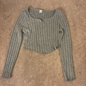 Grå ribbad croppad tröja från Shein - Grå croppad tröja från Shein Teen med ribbad struktur och långärmad design. Tröjan har en snygg v-ringad hals och vågig kant nedtill som ger en cool touch. Perfekt för dig som gillar trendiga och bekväma plagg.