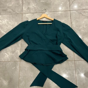Mörkgrön omlottblus från Trendyol - Snygg mörkgrön omlottblus från Trendyol med v-ringning och långa ärmar. Blusen har ett separat brett bälte som kan valfritt knytas i midjan. Materialet är mjukt och stretchigt. Den är helt oanvänd!