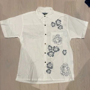 Vit blommig skjorta från Moorea - Snygg vit skjorta frän Flysurf med korta ärmar och ett stilrent blommönster i blätt. Skjortan har bruna trä-knappar framtill och en krage. Skulle behöva en strykning annats helt felfri. Blommigt tryck bäde fram och bak. Riktigt somrig skjorta