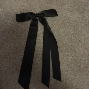 Svart diademrosett i satin - Snygg svart rosett till håret med klämma på baksidan. Gjord i glansigt satinliknande material och har långa band som hänger ner. Perfekt accessoar för att ge en extra touch till din frisyr.