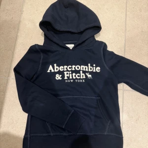 Mörkblå hoodie Abercrombie & Fitch S - Mörkblå hoodie från Abercrombie & Fitch med stor vit logga framtill. Klassisk känguruficka och huva, perfekt för chill dagar. Mjuk insida från Soft A&F Collection. Snygg och enkel att matcha med jeans eller joggers.