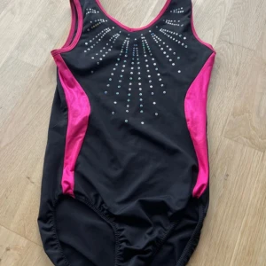 Svart och rosa gymnastikdräkt med diamanter - Snygg svart gymnastikdräkt med starkt rosa detaljer längs sidor och kanter. Framsidan är dekorerad med glittrande stenar i ett strålande mönster. Klassisk sportig modell med breda axelband och rundad hals. 
