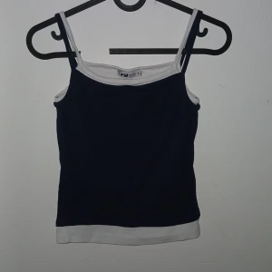 Marinblå och vit linnetopp F8 Sister - Snygg marinblå linnetopp från F8 Sister med vita detaljer längs axelband, halsringning och nederkant. Enkel och stilren design med smala axelband, perfekt för varma dagar eller lager på lager.