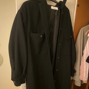 Svart kappa från Ellos, stl 38/40 - Svart overshirt från Ellos i storlek 38/40. Jackan har en rak och loose passform, långa ärmar med knapp vid mudden, två stora bröstfickor med lock och knappar samt hel knäppning framtill.